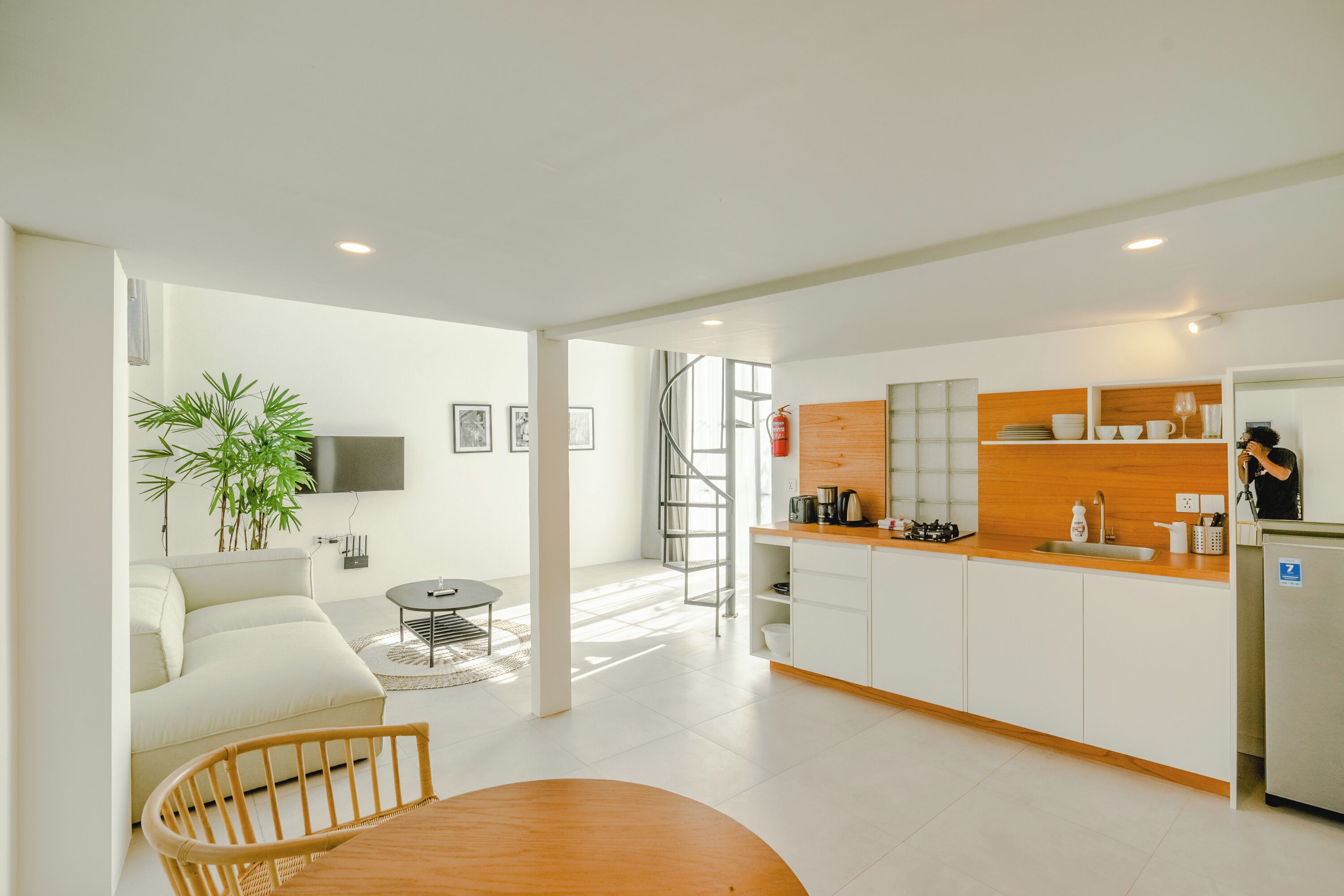 A modern kitchen with white cabinets, a wooden countertop, and a stainless steel refrigerator. There is a dining area with a wooden table and chairs, and a living room with a sofa and a TV.