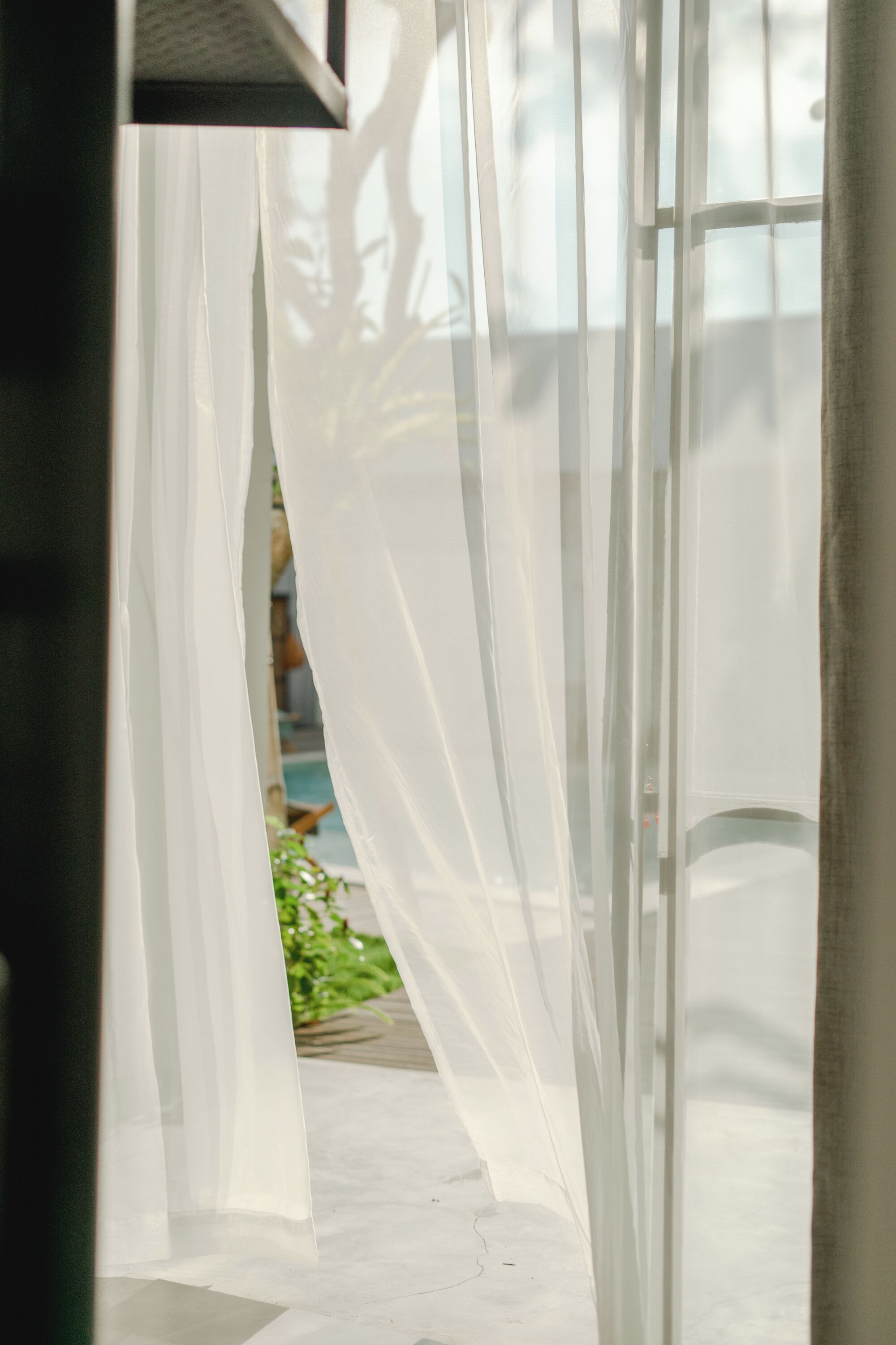 Vista a través de cortinas blancas y traslúcidas hacia un cuarto con piso de concreto y un vistazo de vegetación en el exterior.