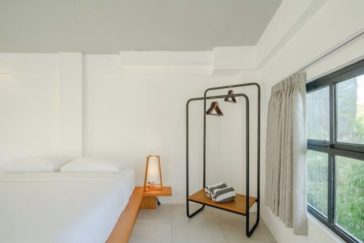 A minimalist hotel room with a bed, a small wooden side table, a wall-mounted hanging chair, and a large window with curtains.