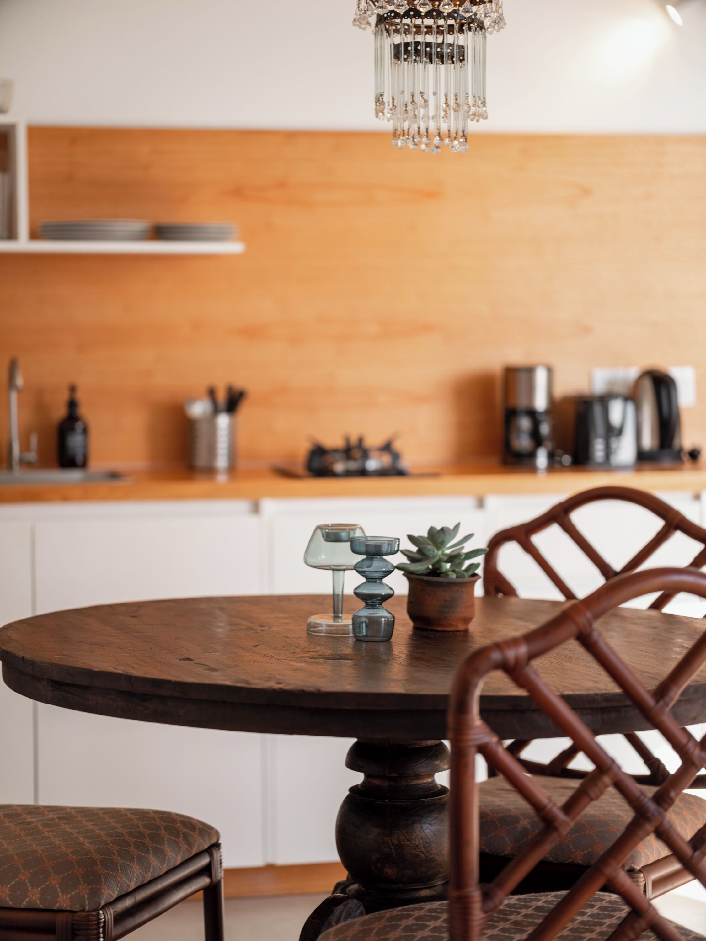 Une table de salle à manger ronde en bois, avec une chaise placée au premier plan. À l’arrière-plan, on aperçoit un coin cuisine avec un plan de travail en bois, une machine à café et une plante en pot.