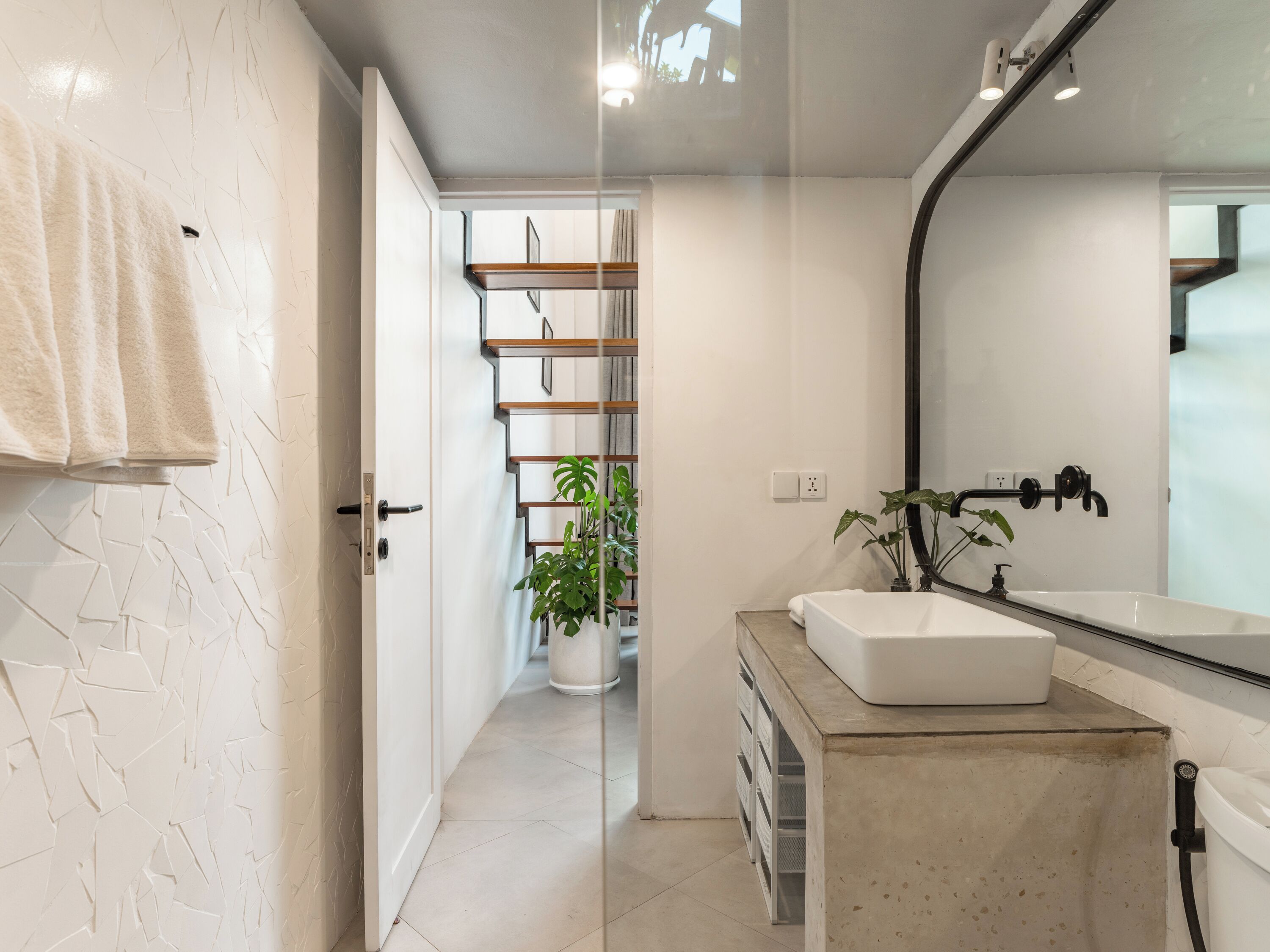 Un baño moderno con un lavamanos blanco, un espejo y una ducha. Hay un perchero a la izquierda y una planta cerca del lavamanos.