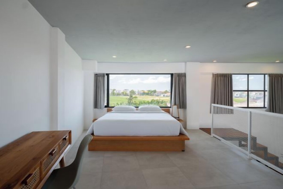 A modern bedroom with a large bed, a wooden desk, and a view of a green landscape through large windows.