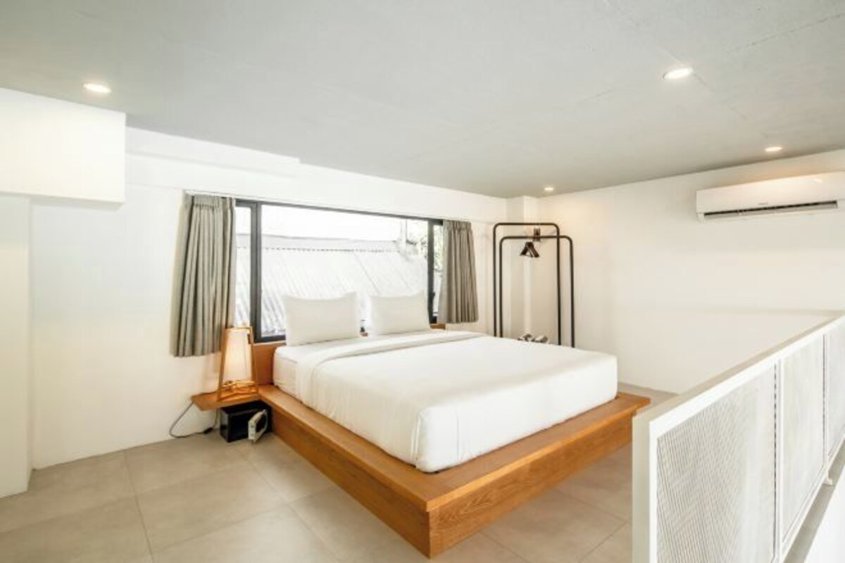 A modern hotel room with a wooden platform bed, a bedside lamp, and a view through a large window.