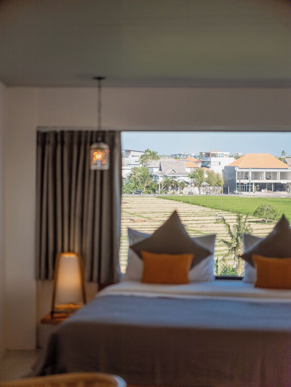 A hotel room with a bed, a nightstand, a lamp, and a view of a building and fields. - AKA Tropicana Loft Canggu (Canggu)