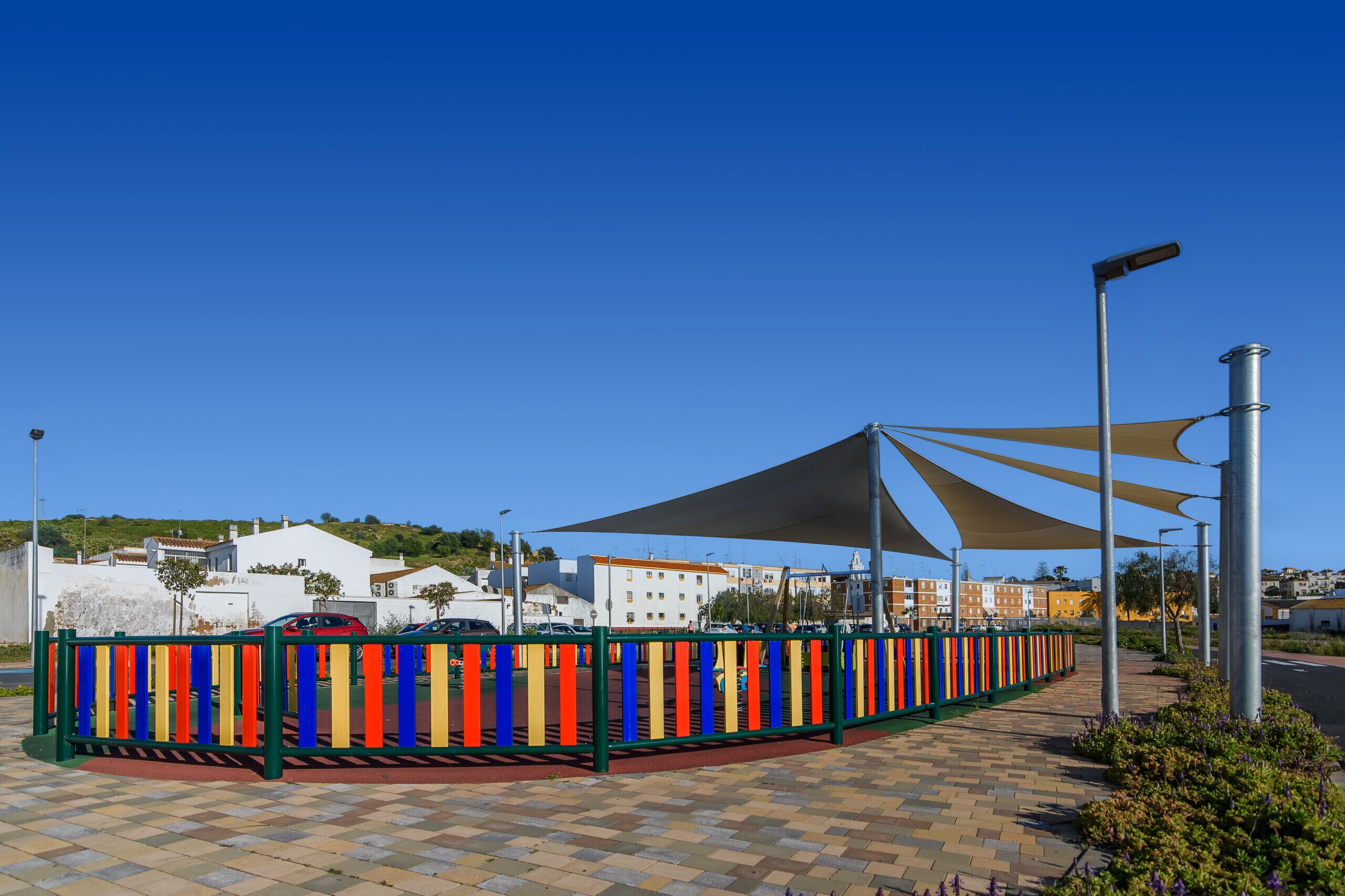 Un sendero pavimentado con una valla colorida, una zona con sombra y edificios al fondo.