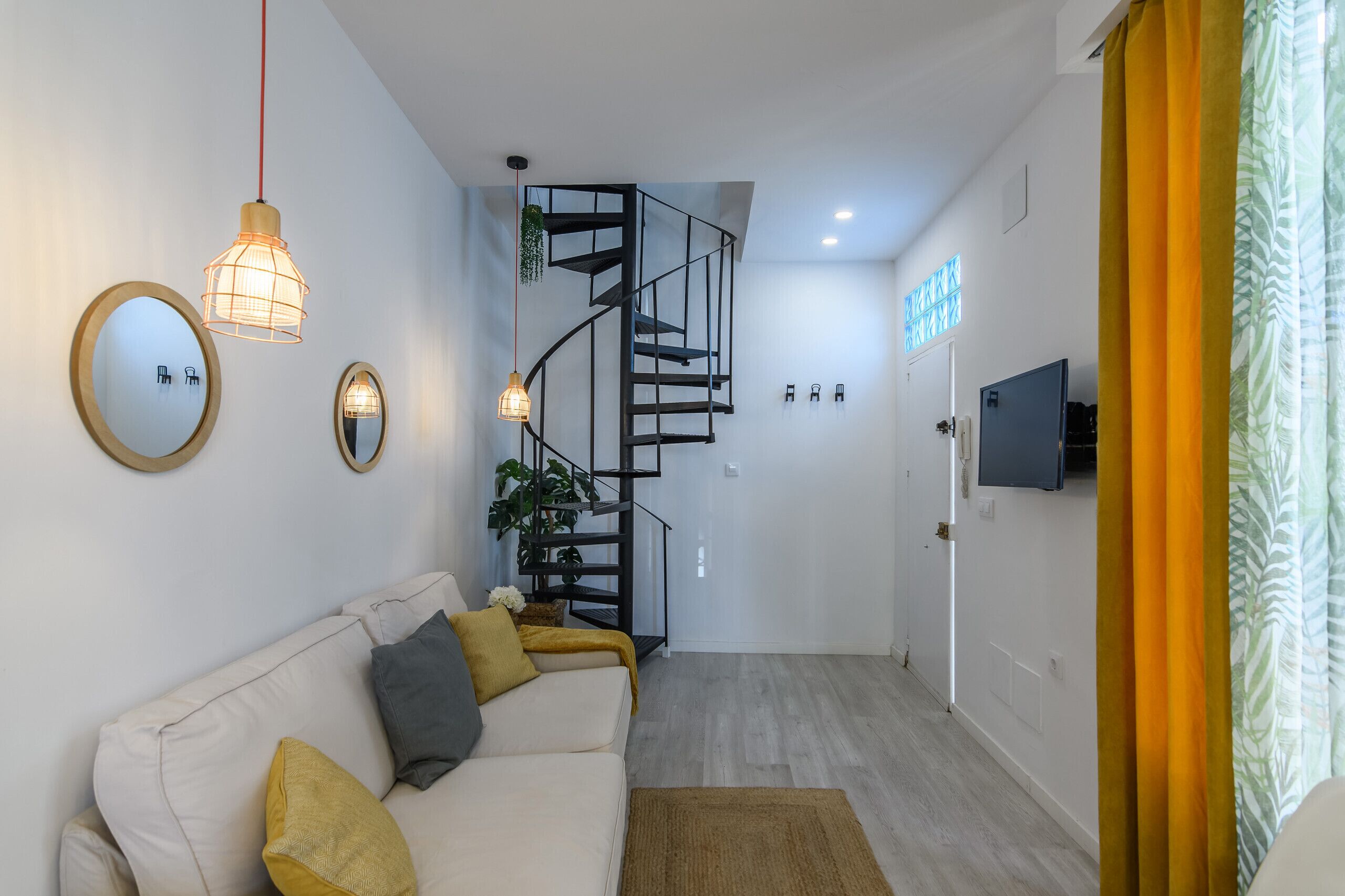 Una sala de estar moderna con un sofá, dos luces fijadas a la pared, una escalera en espiral y un televisor en la pared.
