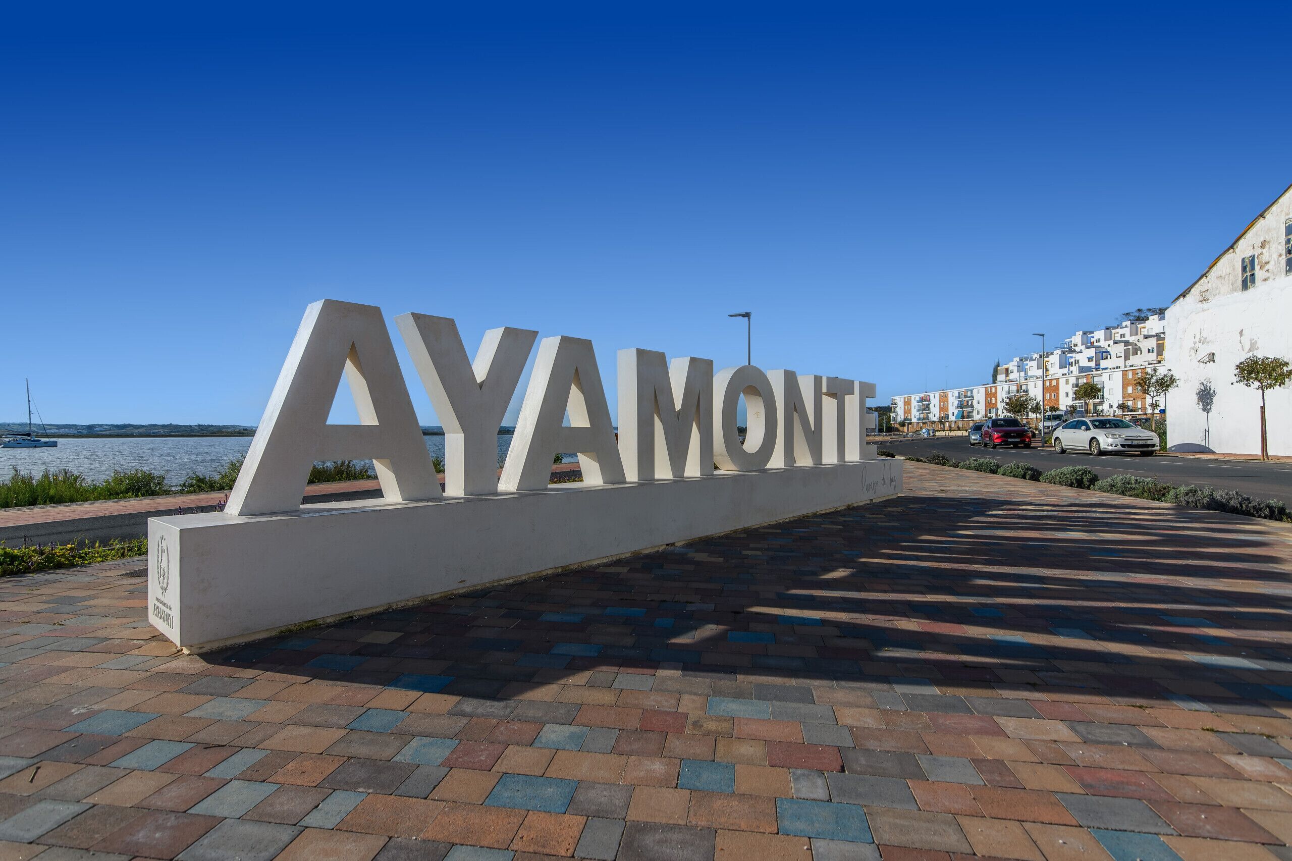 Un letrero grande con la palabra "AYAMONT" en letras blancas sobre una zona pavimentada, con vista a una masa de agua y edificios al fondo.