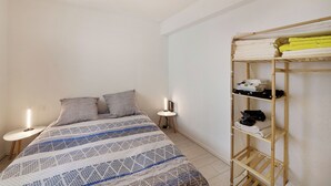 A bedroom with a bed, pillows, a bedside table with a lamp, and a wooden shelf with folded towels. - Le Petit Coin de Lavaur (Lavaur)