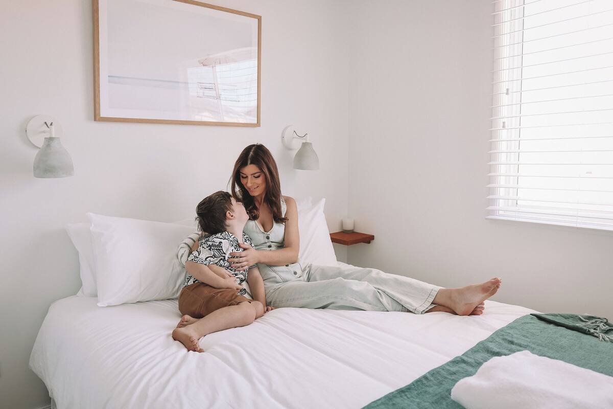 Un dormitorio con cama, mesita de noche, lámpara de pared y ventana con persianas.