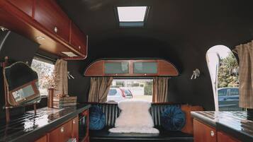 A compact RV interior with a kitchenette, a small seating area with a fur throw, and a window with curtains.