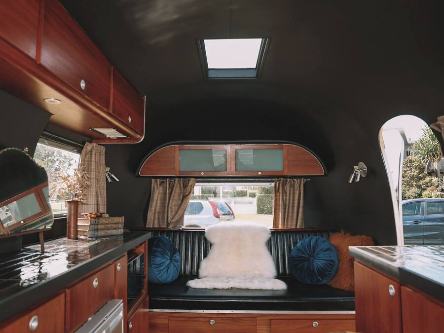 A compact RV interior with a kitchenette, a small seating area with a fur throw, and a window with curtains.