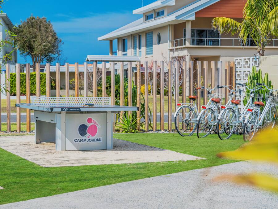 Une table de billard ornée du logo « CAMP JORDAN », entourée d’une zone délimitée par une clôture, avec des vélos garés à proximité.