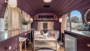Una habitación compacta y bien iluminada con una cortina a cuadros, una pequeña mesa con un florero, dos sillas acolchadas, una estantería con plantas y un refrigerador.