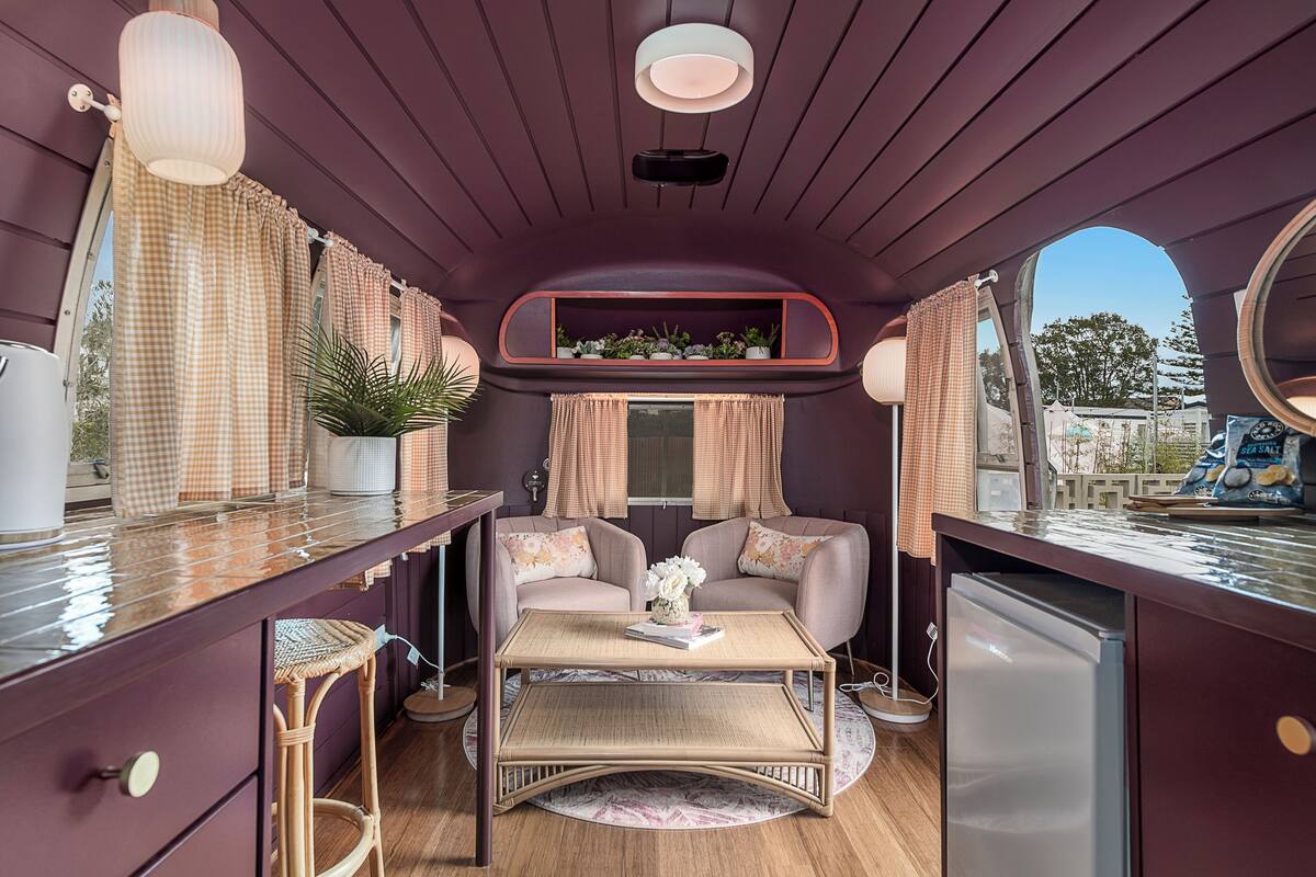 Una habitación compacta y bien iluminada con una cortina a cuadros, una pequeña mesa con un florero, dos sillas acolchadas, una estantería con plantas y un refrigerador.
