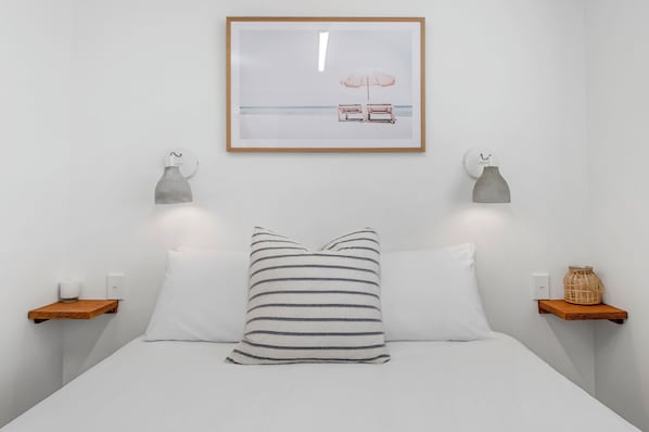 A neatly arranged bedroom with a bed, striped pillows, a framed picture, bedside tables with lamps, and a small basket on a shelf. - Camp Jordans (Port Macquarie)
