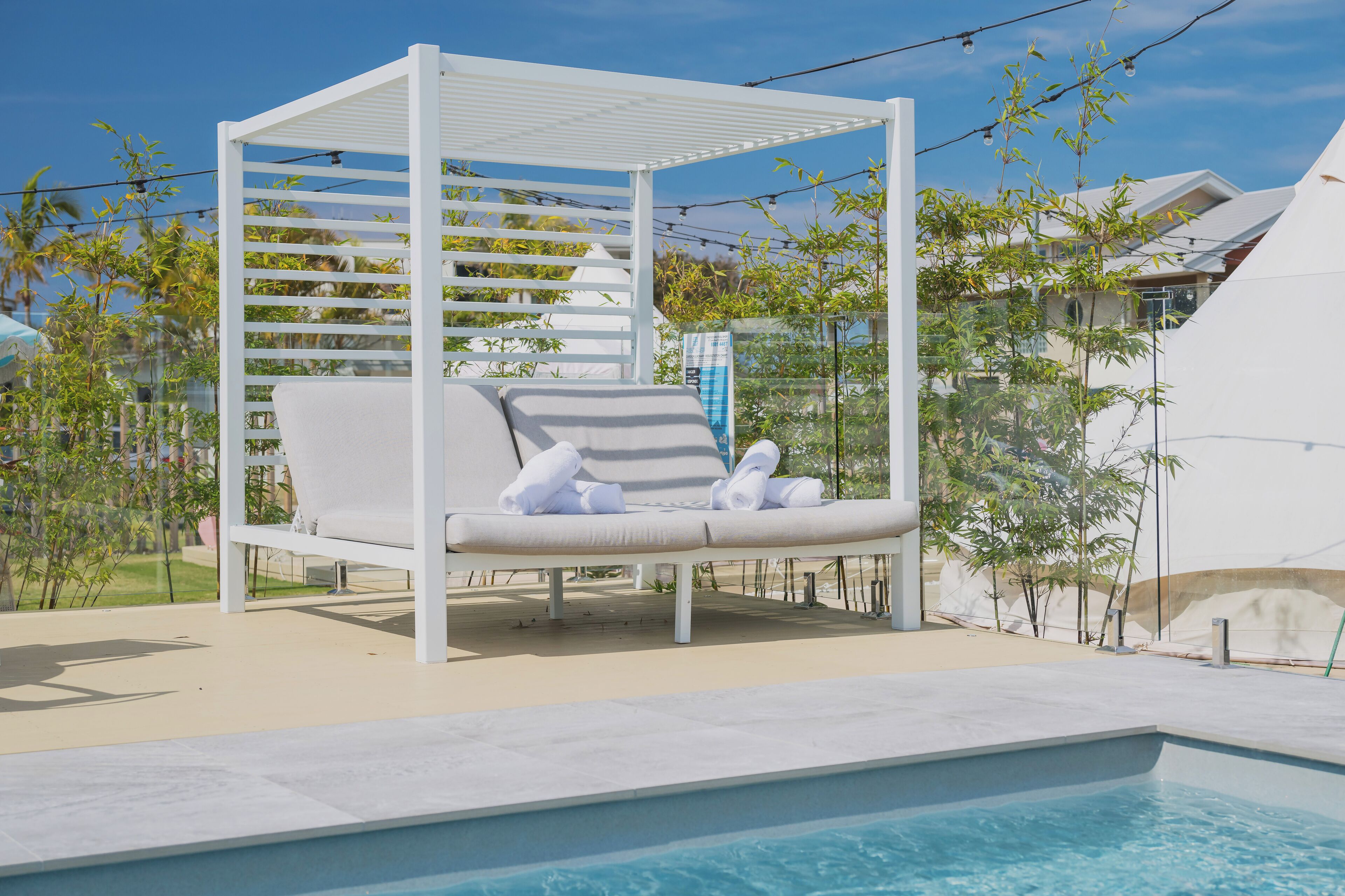 Un área junto a la piscina con una pérgola blanca, una tumbona con cojín a rayas y una piscina.