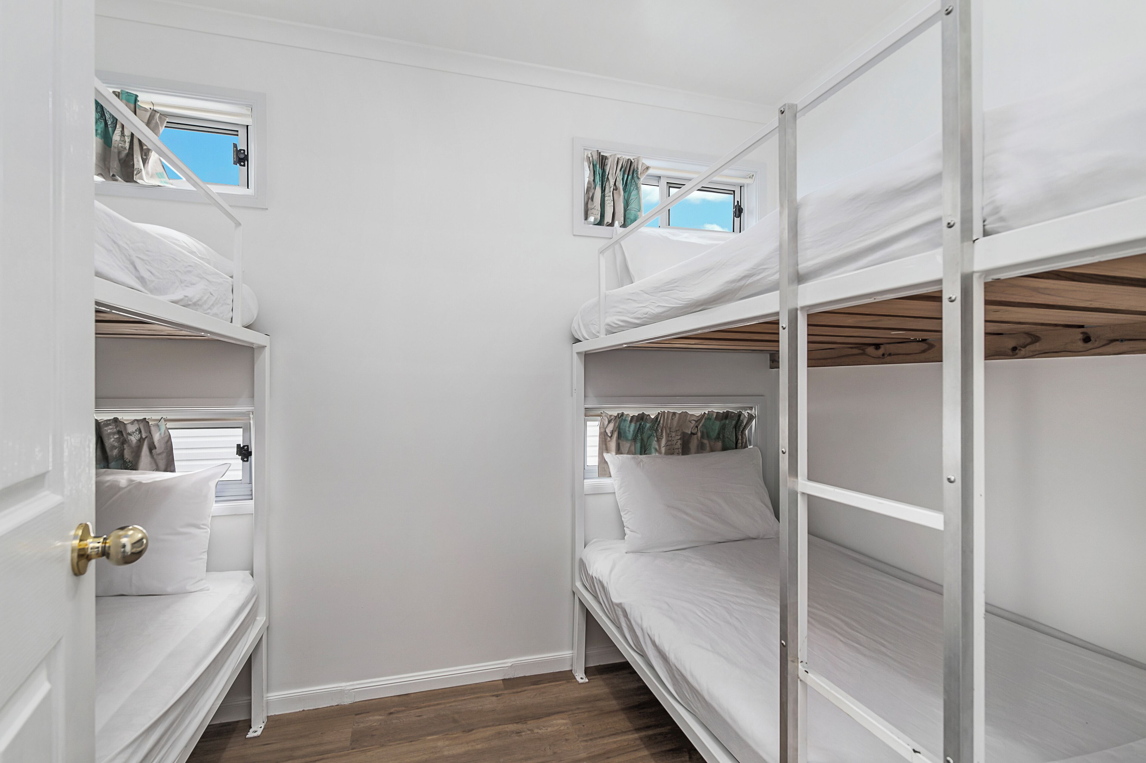 A room with three bunk beds, each with a window. The room has white walls and wooden flooring.