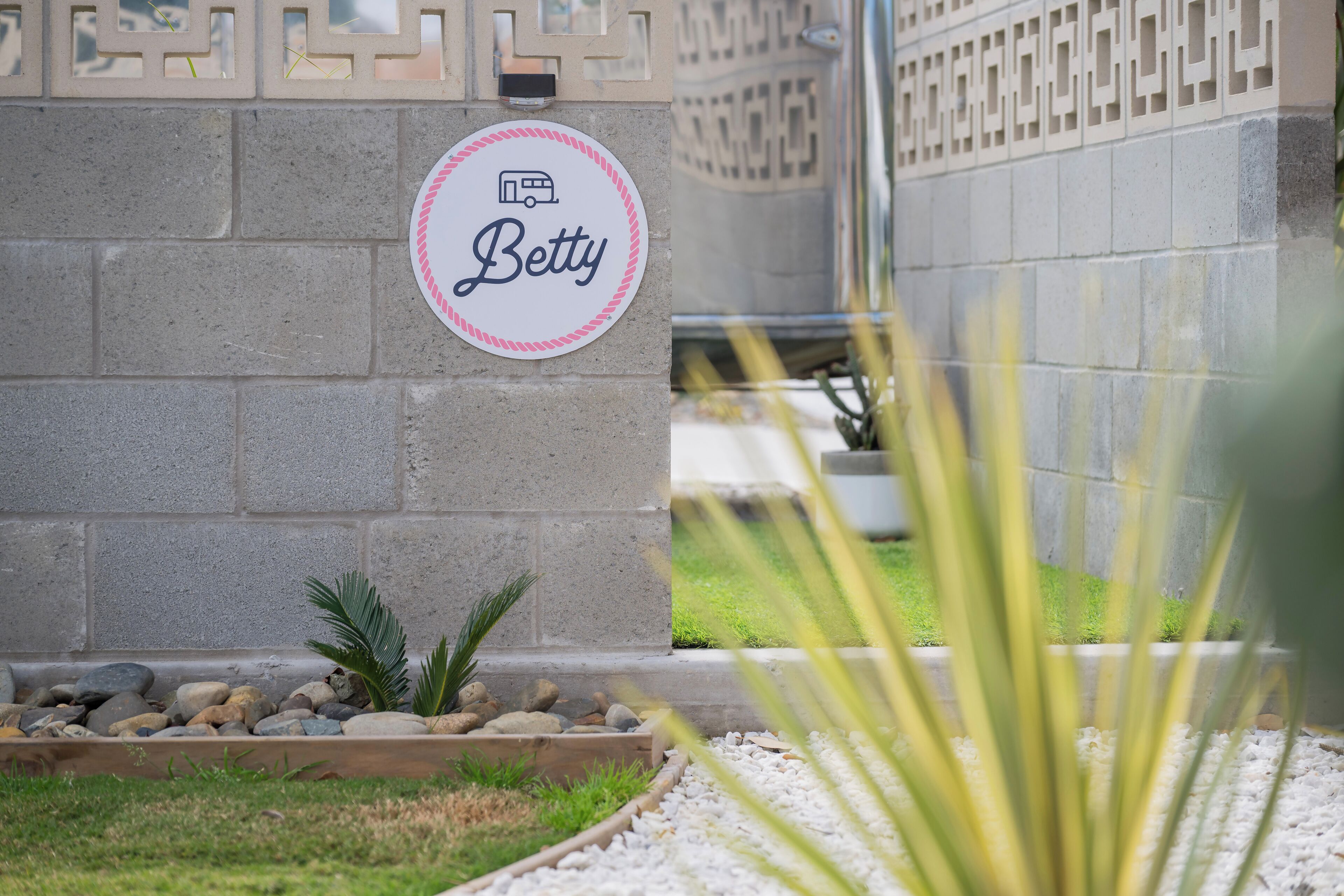 Une enseigne portant le nom « Betty » est fixée à un mur, et elle est entourée d’un jardin agrémenté de rochers et de plantes.