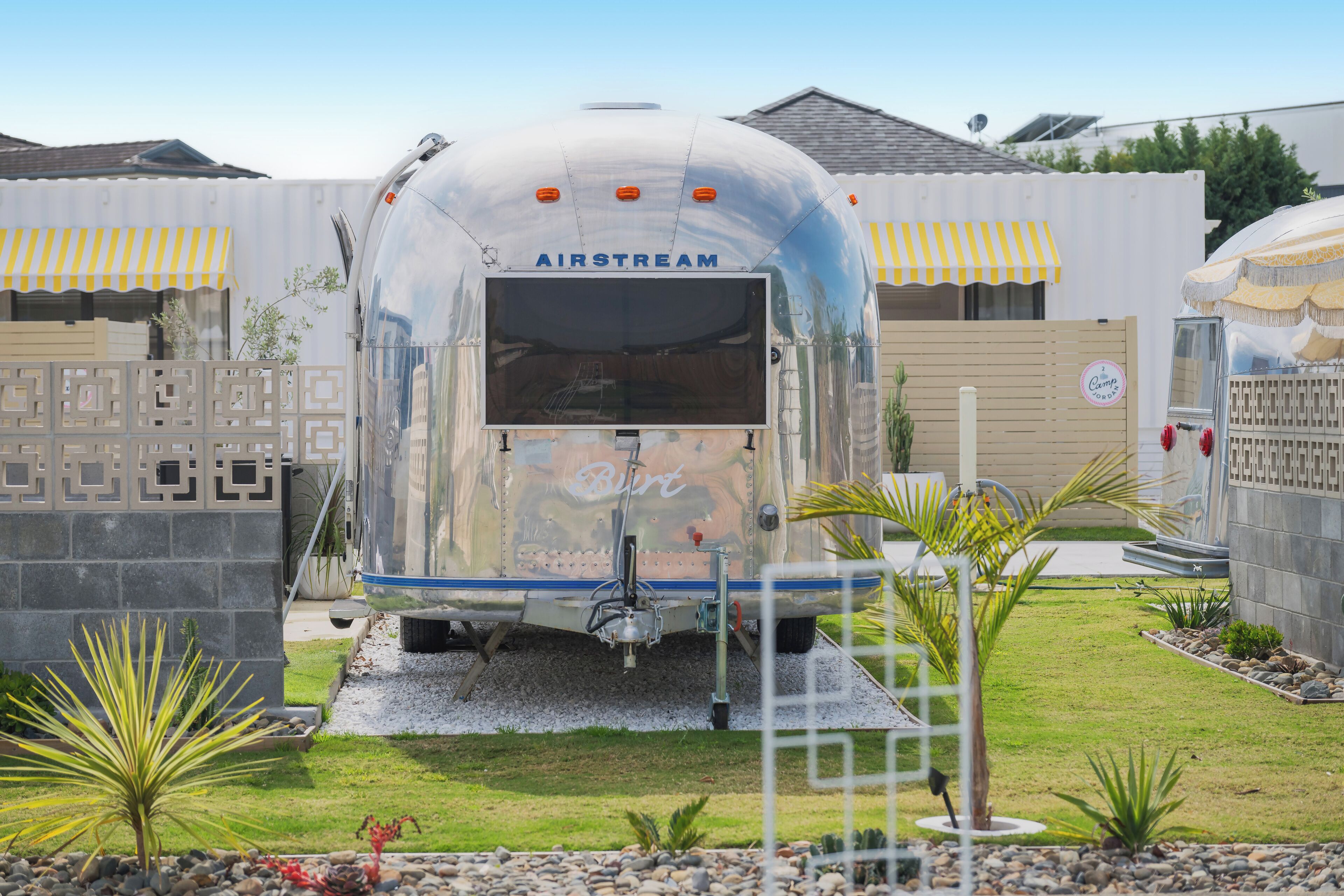 Une caravane Airstream ancienne, ornée du logo « Birt », entourée d’un jardin aménagé avec des palmiers et une clôture décorative.