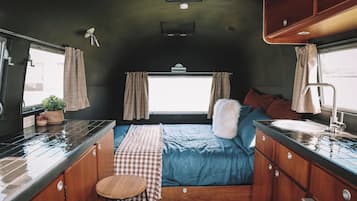 Un intérieur de camping-car compact comprenant un coin cuisine, un lit avec une literie bleue et une fenêtre avec des rideaux.