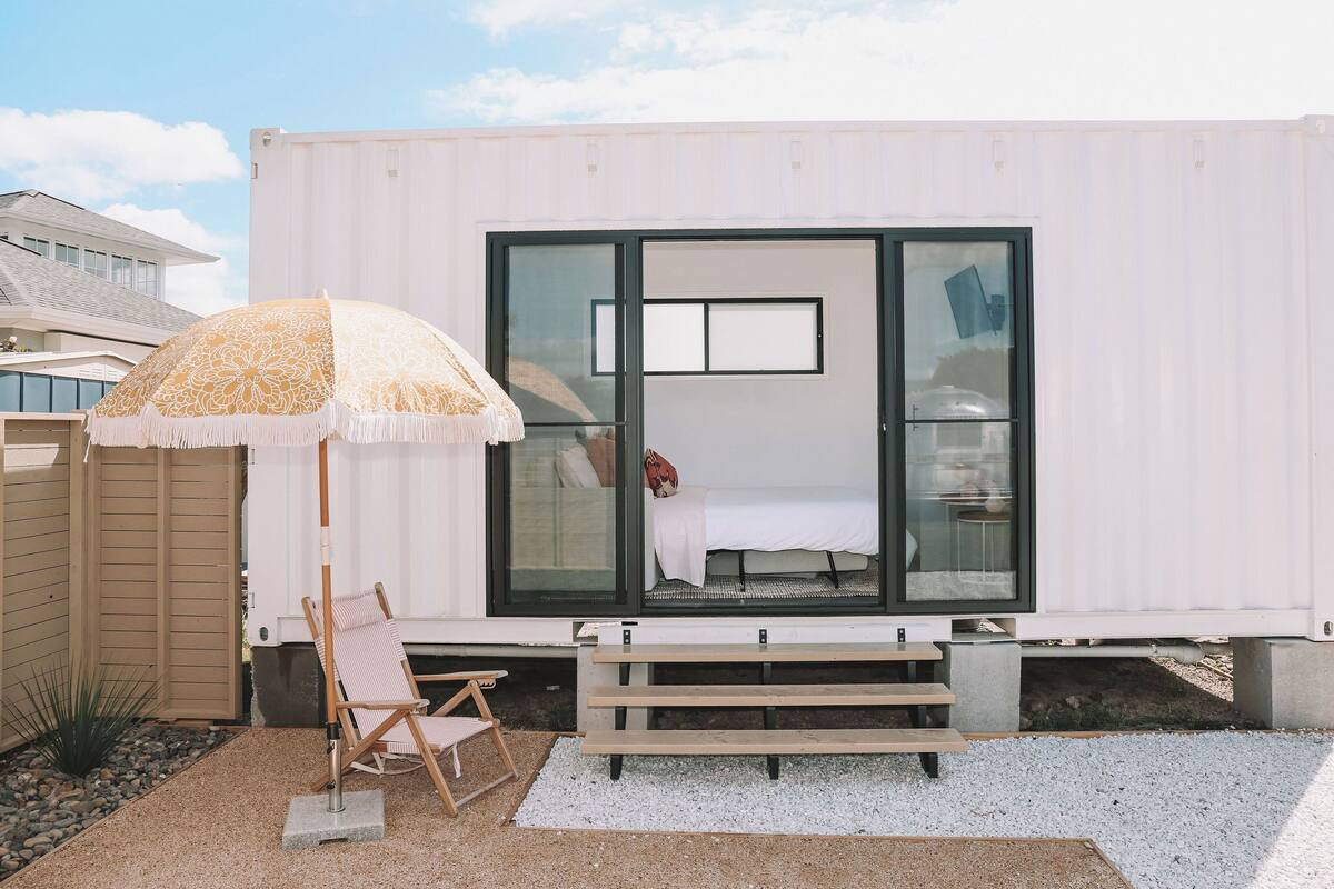 Una vivienda moderna tipo contenedor con un ventanal grande, una banca y un paraguas.