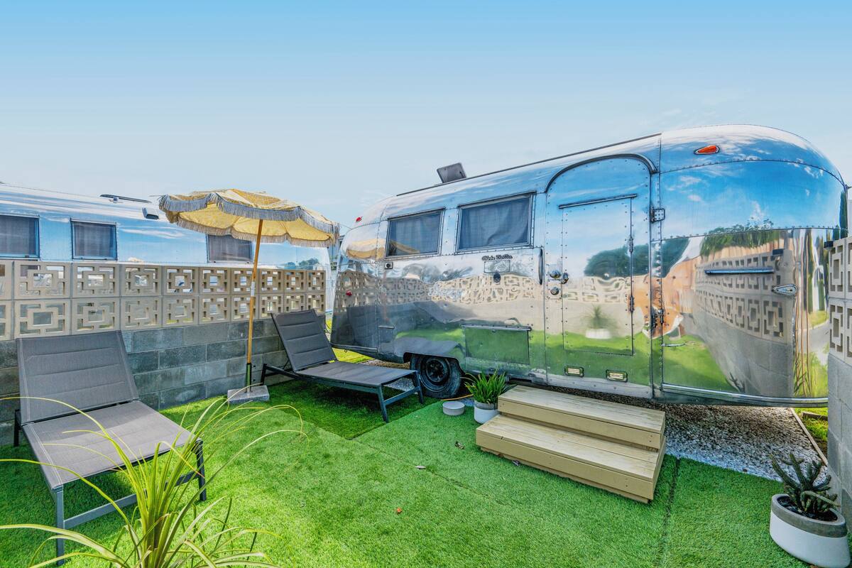 Un remolque con superficie reflectante, una pequeña terraza, una silla de descanso, una mesa y una planta en maceta.