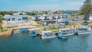 Un puerto deportivo con varias embarcaciones amarradas, un pequeño edificio con un letrero y una hilera de casas al fondo.