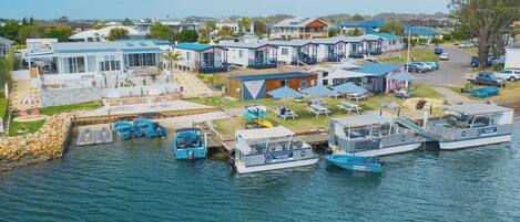 A marina with several boats docked, a small building with a sign, and a row of houses in the background.