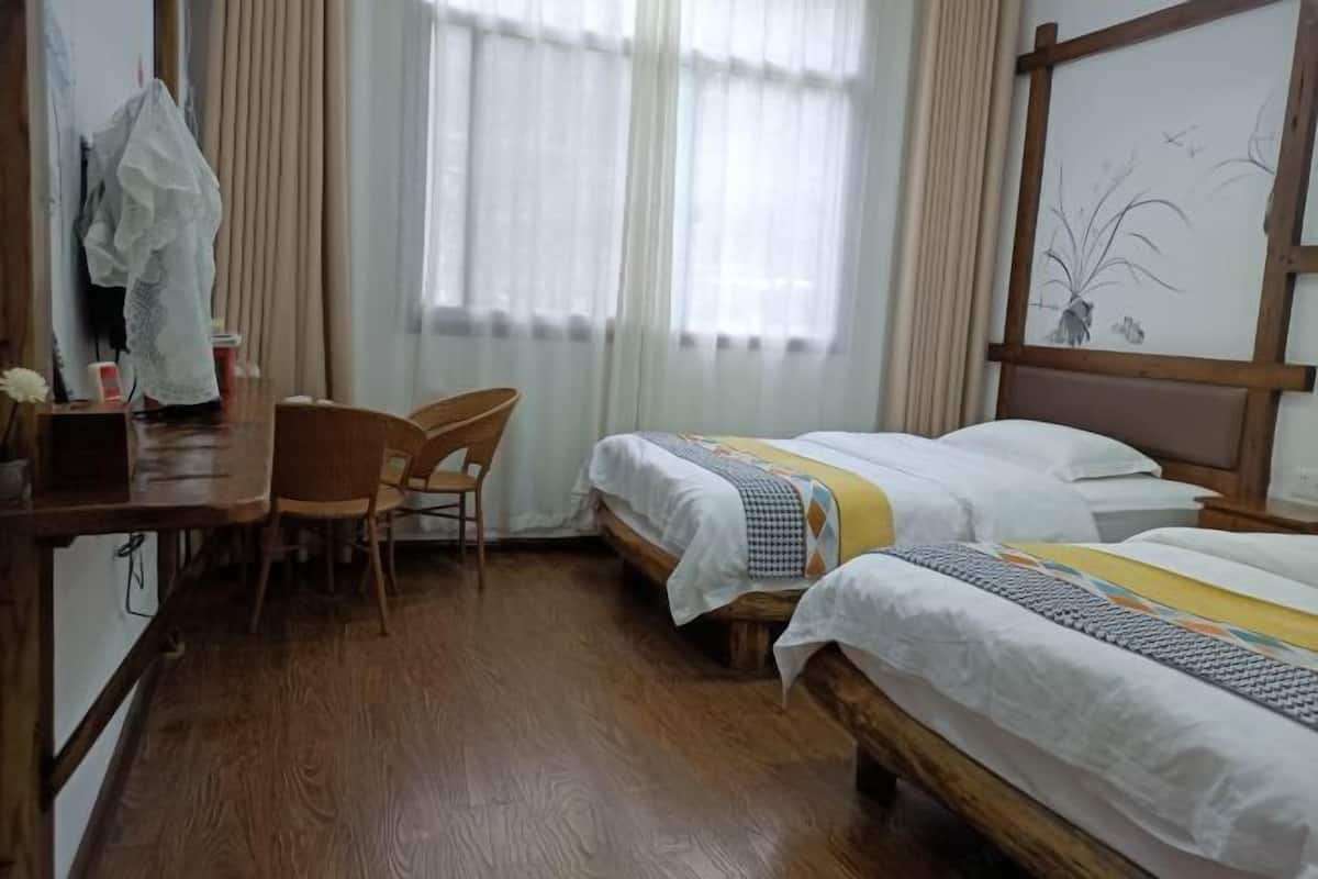 A hotel room with two single beds, a wooden desk, a chair, and a large window with sheer curtains.