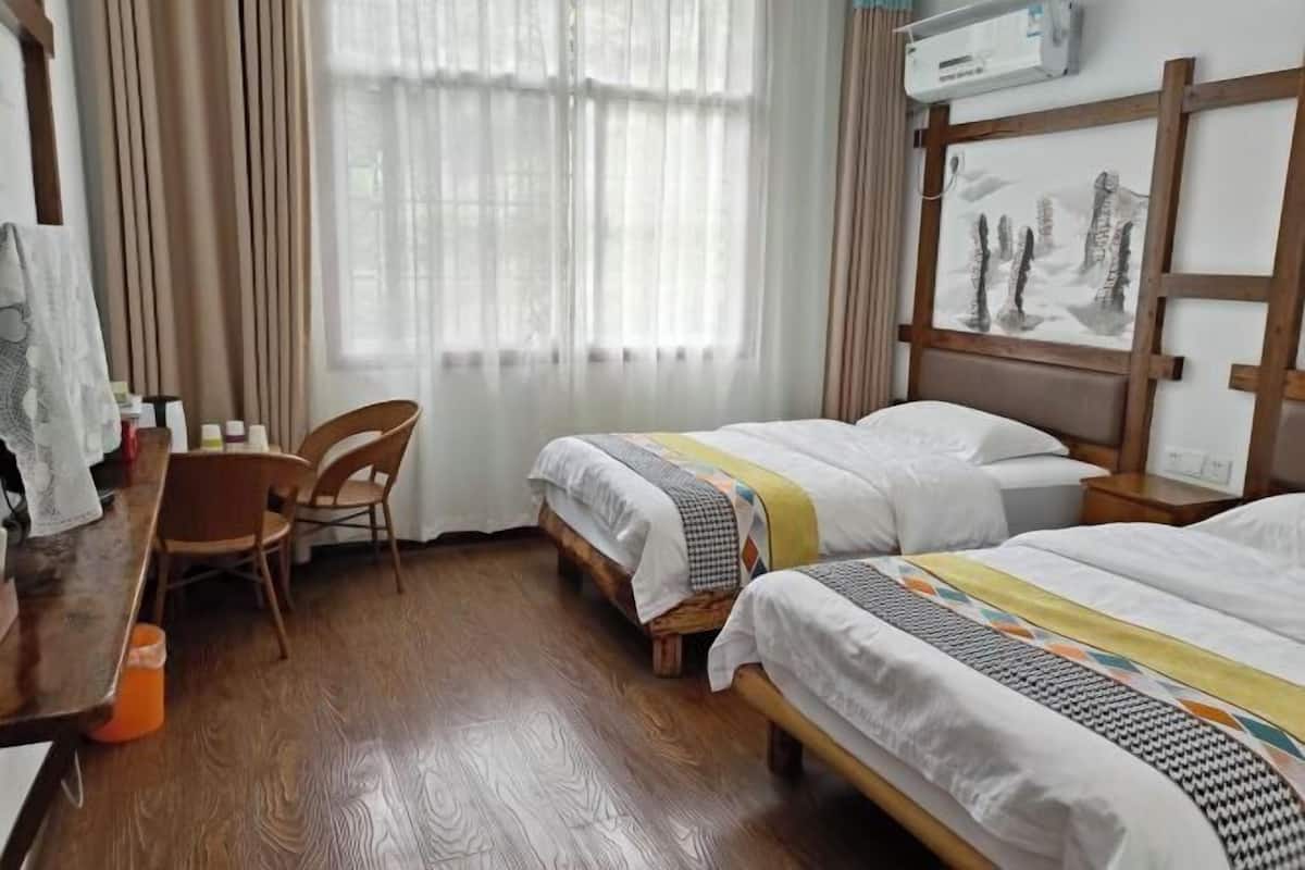 A hotel room with two beds, a wooden floor, a desk with a chair, and a window with curtains.