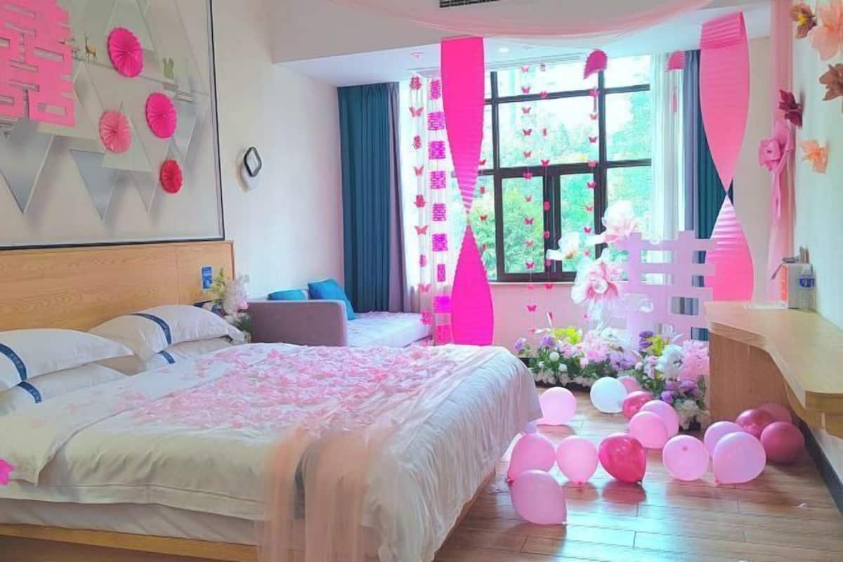 Un dormitorio con cama, almohadas, una mesita de noche, una ventana con cortinas y una pared decorada.