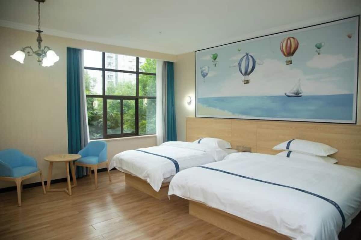 Habitación de hotel con dos camas, una mesita pequeña, dos sillas, una araña, un ventanal grande con cortinas y un cuadro de globos aerostáticos.