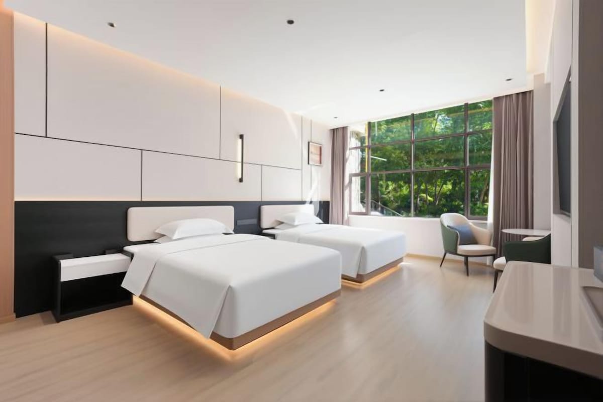 A modern hotel room with two beds, a desk, and a large window offering a view of greenery.