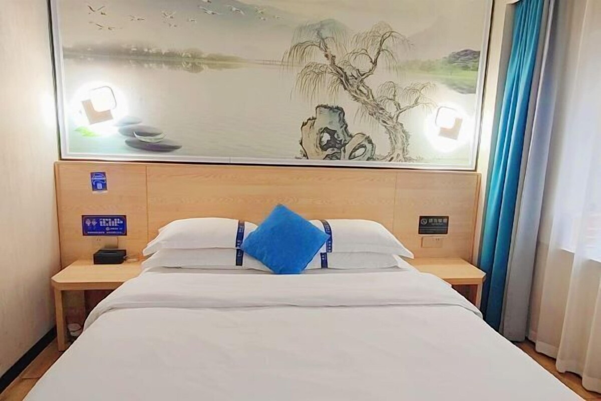 A hotel room with a bed, a nightstand, a wall-mounted artwork, and a window with curtains.