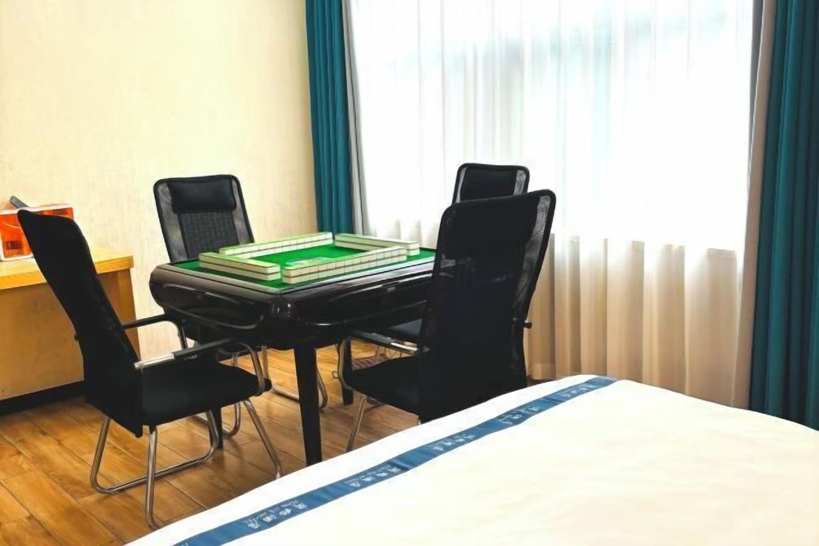 A hotel room with a bed, a table with a green tray, two black chairs, and a window with curtains.