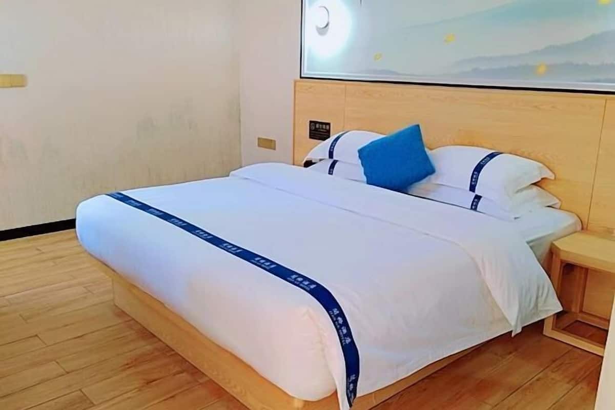 Una habitación de hotel con estructura de cama de madera, ropa de cama blanca, almohada azul, una mesita de noche pequeña y una lámpara fijada a la pared.