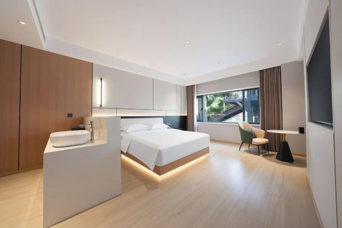A modern hotel room with a large bed, a bedside table, a built-in sink, and a window with a view.