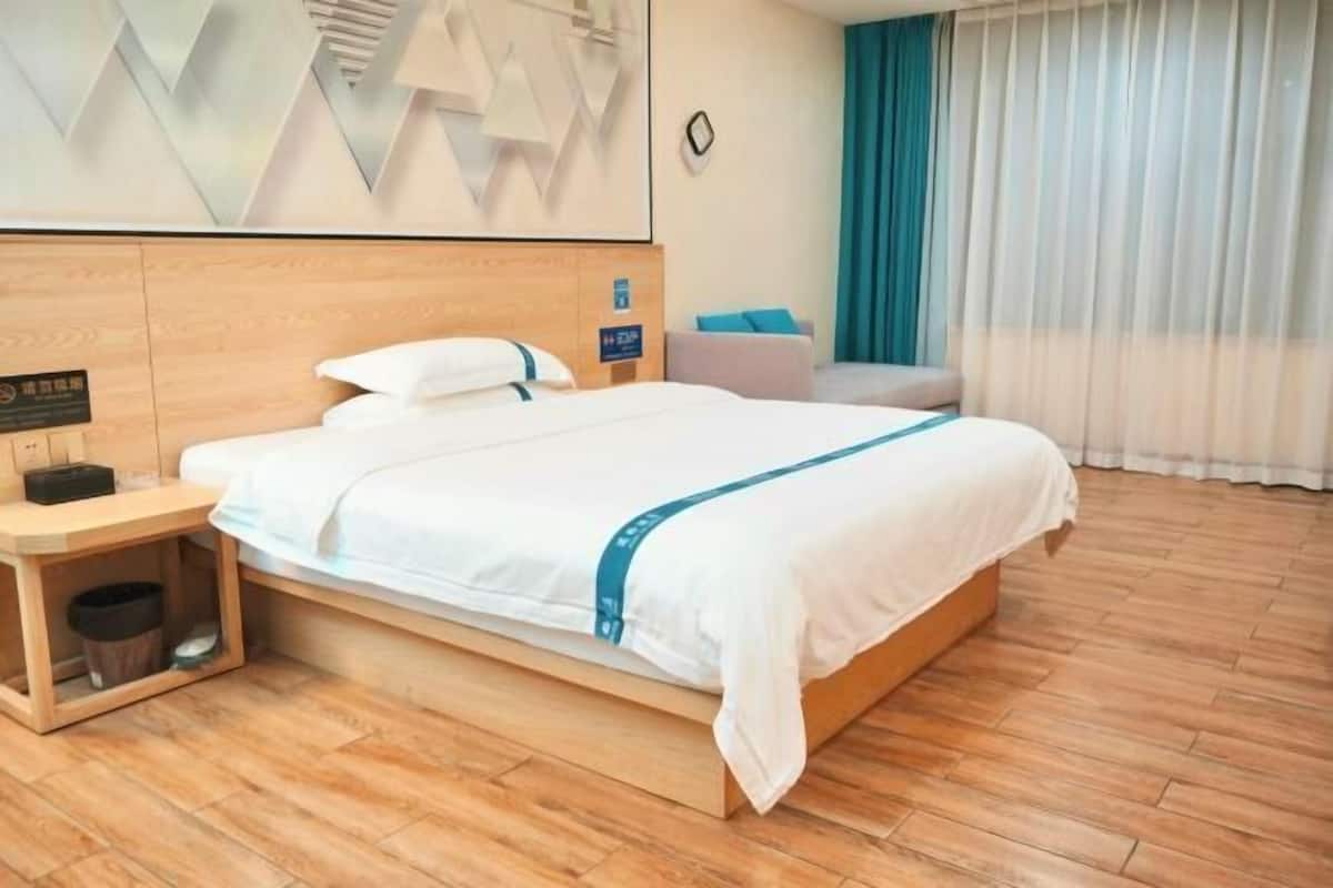 Habitación de hotel con cama de madera, mesita de noche, un cubo pequeño de basura, sofá y un ventanal con cortinas.