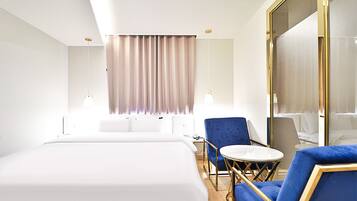 A hotel room with a large bed, a small round table, two blue armchairs, and a large mirror.