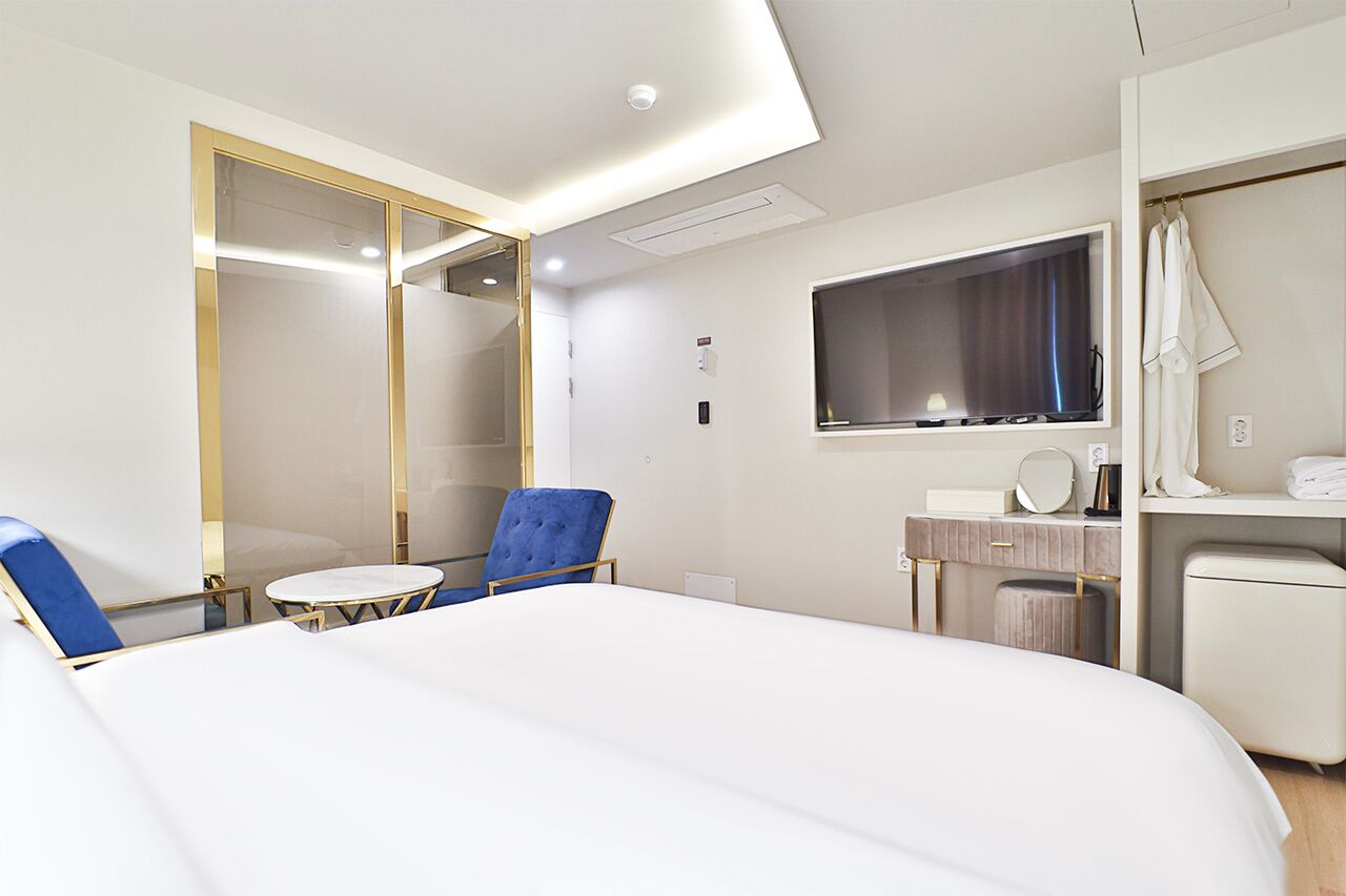 Quarto de hotel moderno com uma cama grande, uma poltrona azul, uma mesa redonda pequena, uma toucador com espelho e uma televisão.