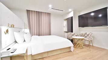 A modern hotel room with a large bed, a bedside table with a remote control, a round table with chairs, a large TV, and a window with curtains.