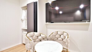 A modern room with a large flat-screen TV mounted on the wall, a round white table with gold legs, and two upholstered chairs with a unique design. - HADAN DENBASTA HOTEL (Busan)