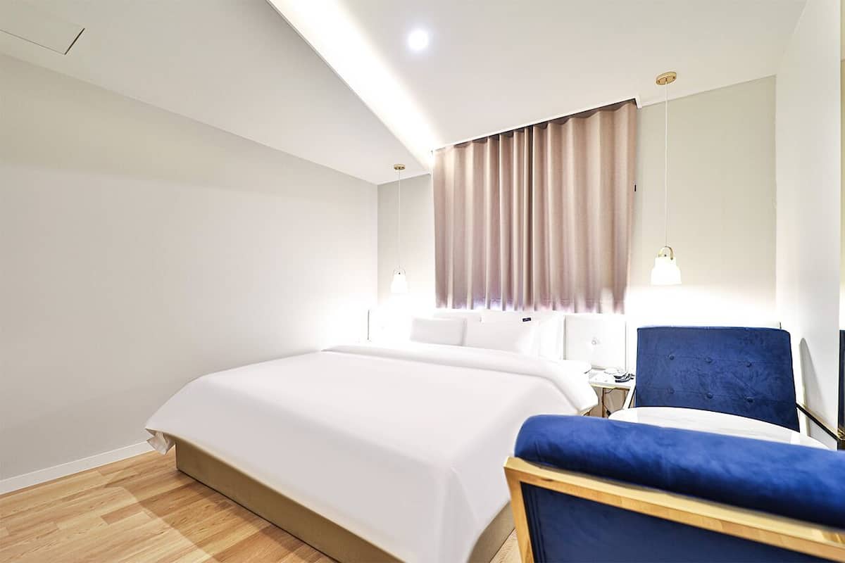 Una habitación de hotel moderna con una cama grande, una silla azul y una mesita de noche con lámpara.