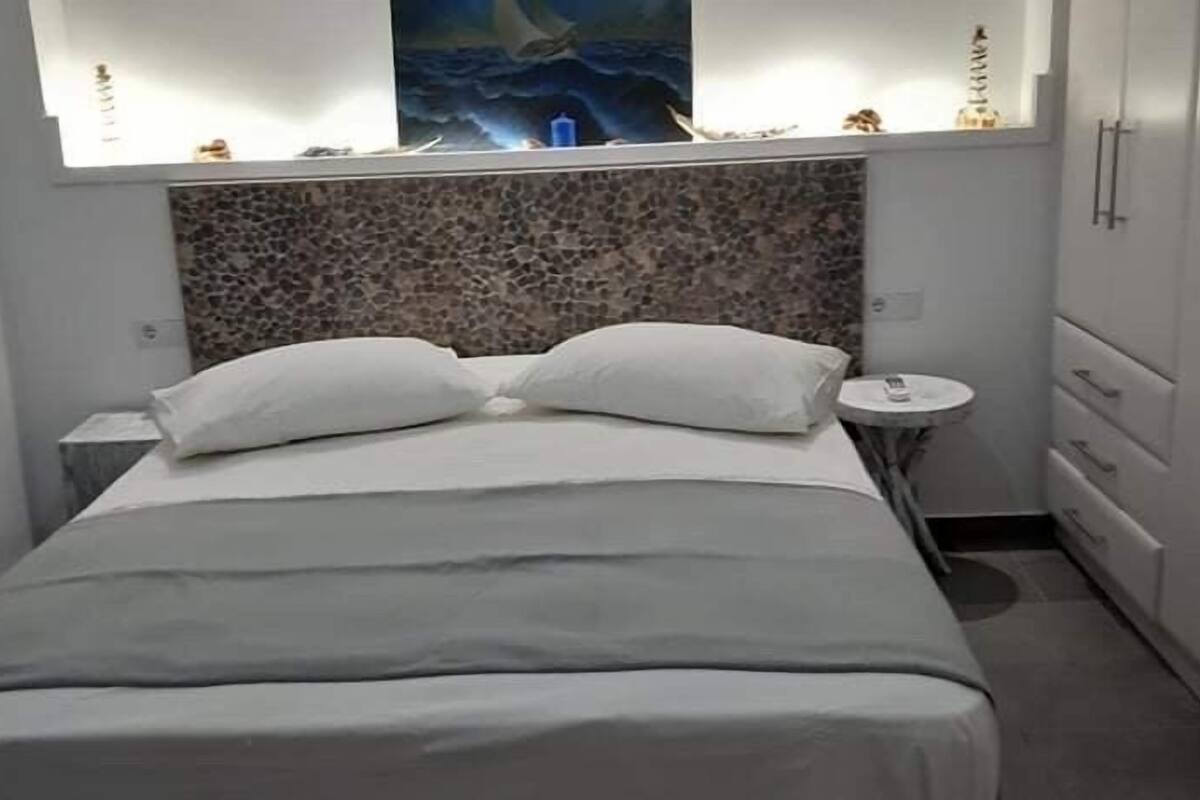 Un dormitorio con cama, cabecera, mesita de noche, una pequeña mesa redonda y un armario.