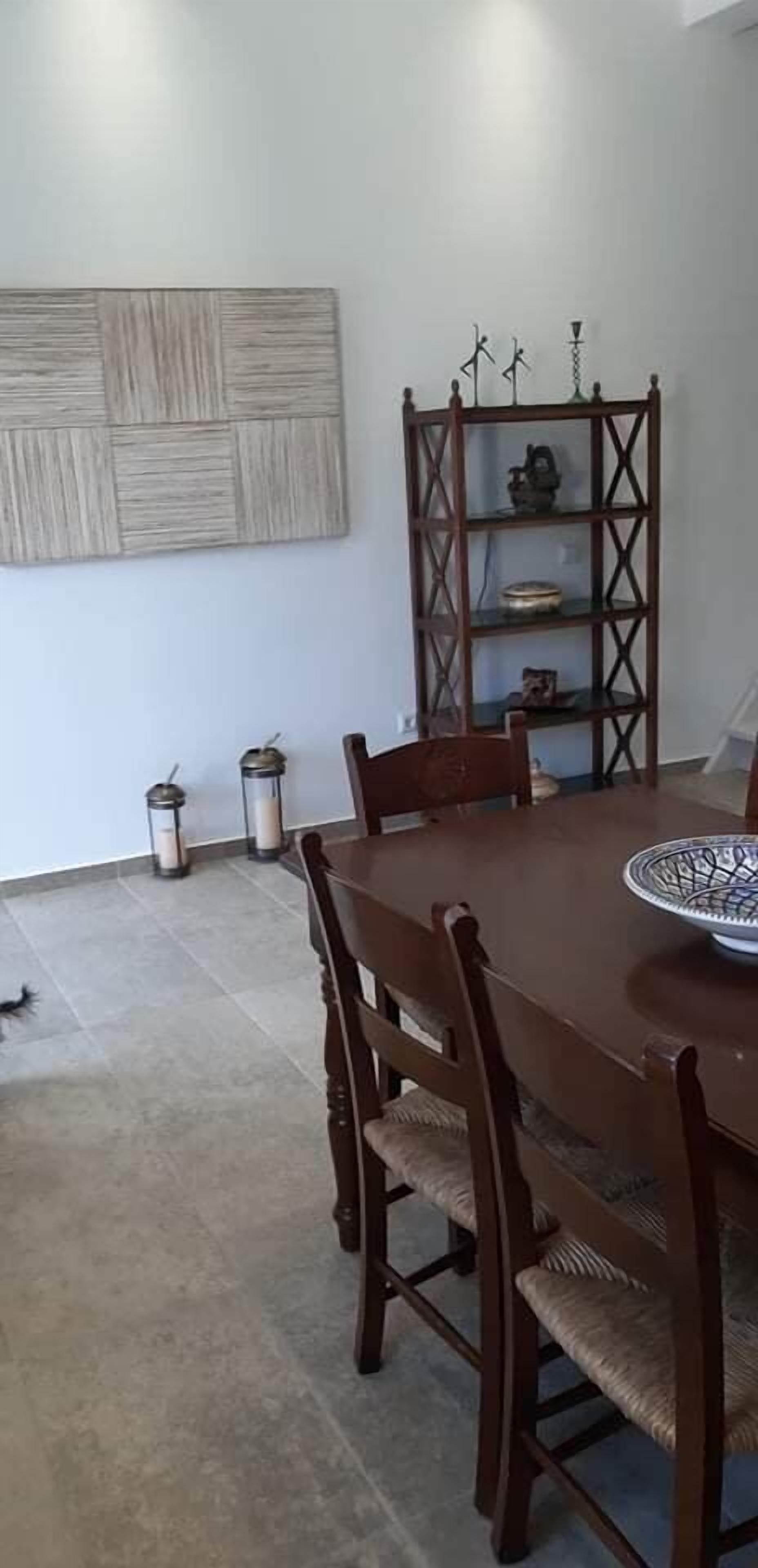 Un comedor con una mesa y sillas de madera, un adorno en la pared y una estantería metálica con varios objetos.