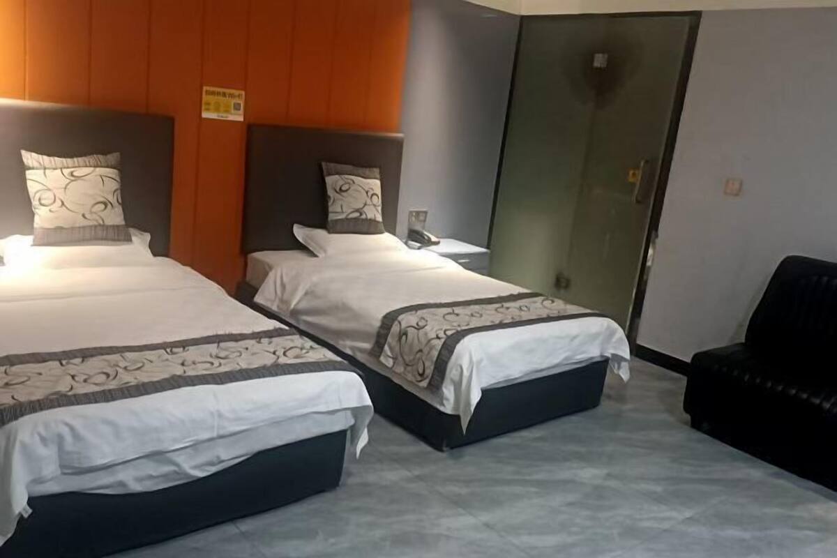 Dos camas individuales con sábanas blancas y almohadones estampados, un sillón de cuero negro y una puerta de vidrio.