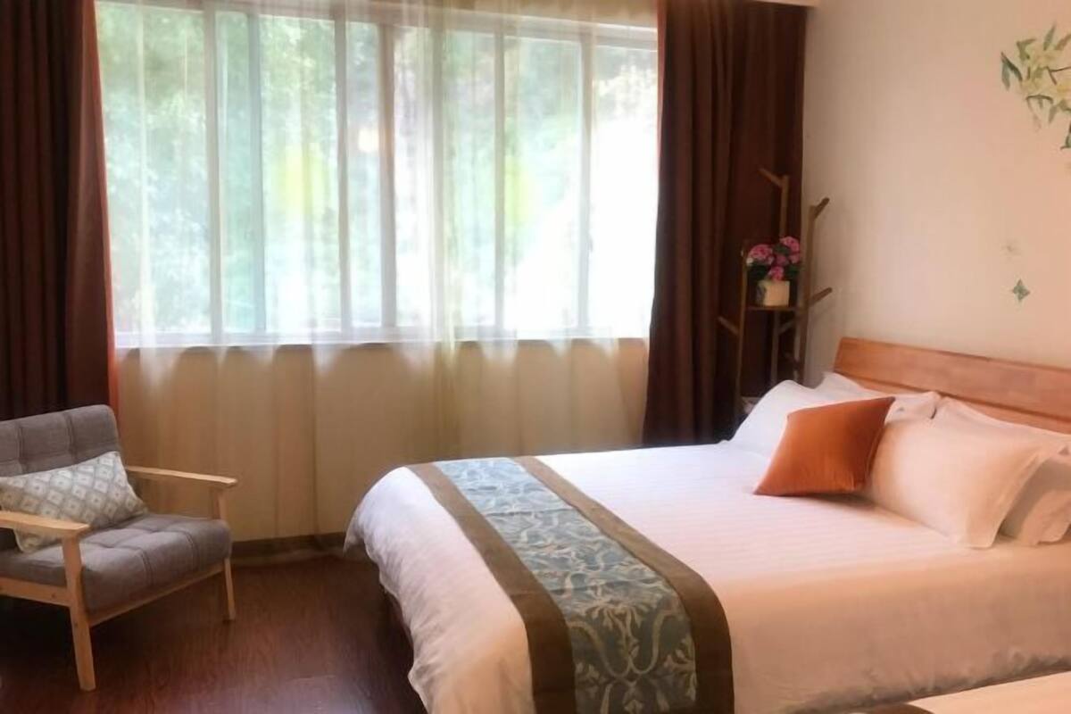 Hotelkamer met een bed, een stoel, een raam met gordijnen en een wandplank met bloemen.