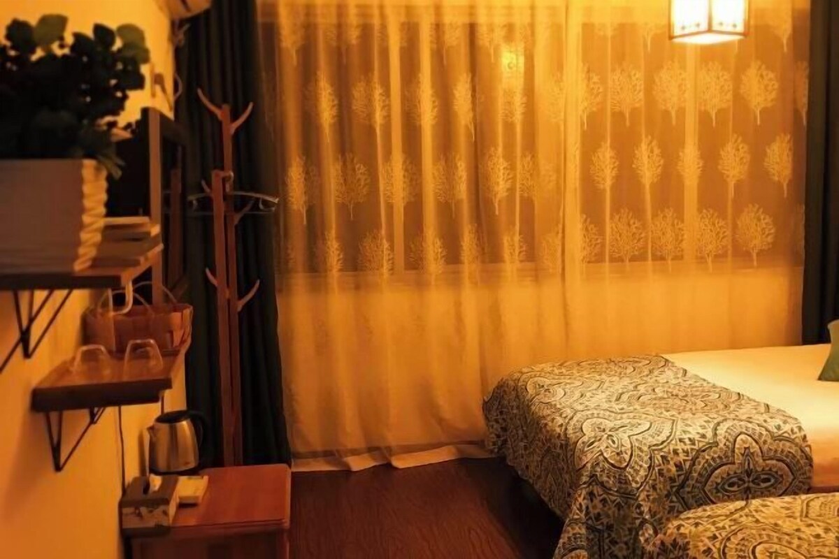 Une chambre d’hôtel avec un lit, une table de chevet, une lampe, une plante et une fenêtre avec des rideaux.
