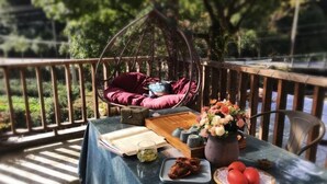 Een houten terras met een hangstoel, een tafel met een boek, een vaas met bloemen en een bord met tomaten.