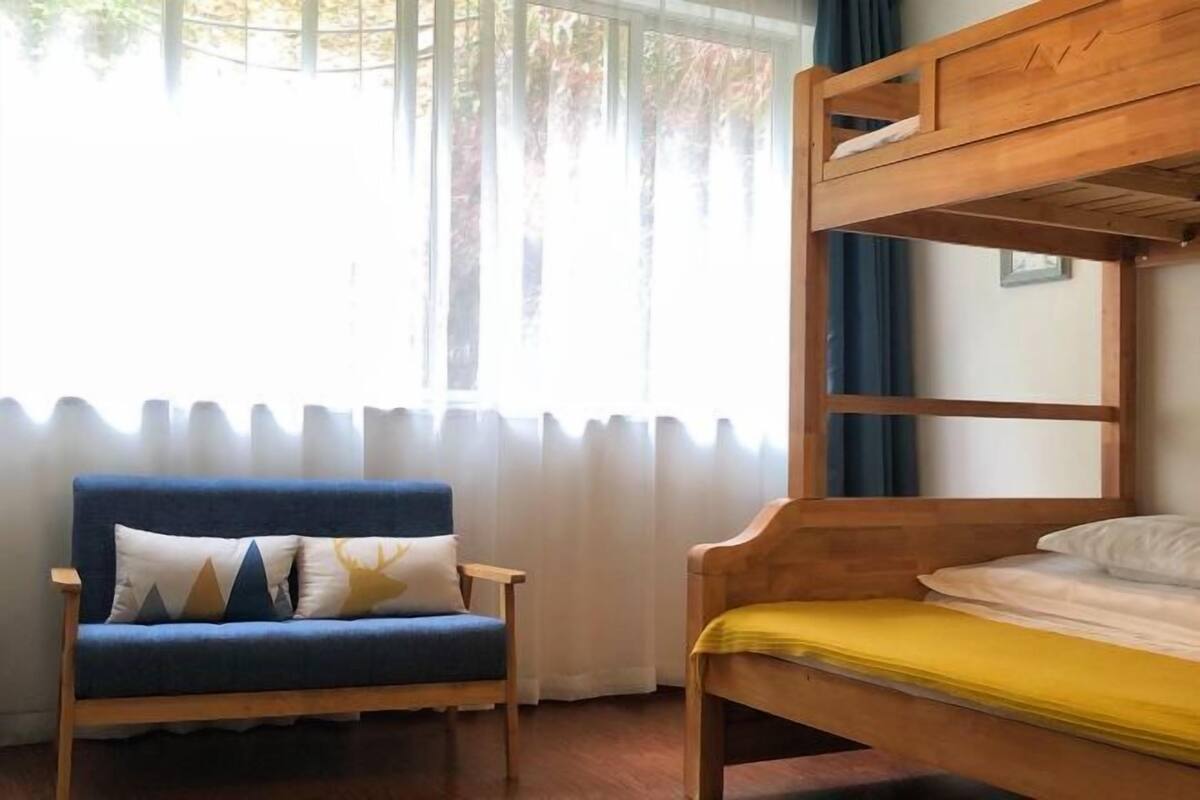 Een kamer met een stapelbed, een blauwe sofa met gekleurde kussens, en een raam met gordijnen.